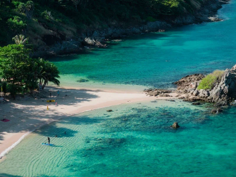 A tropical beach with clear turquoise waters, white sandy shore, palm trees, and rocky cliffs in the background.