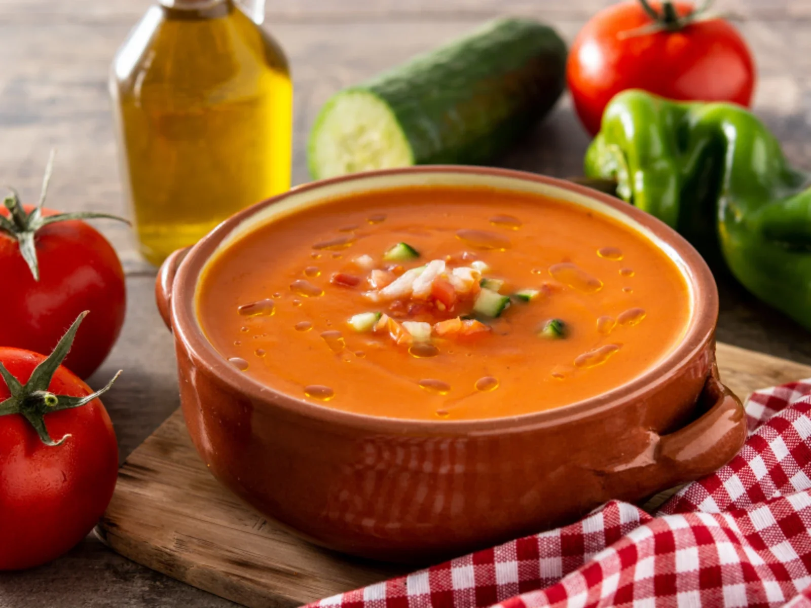 A bowl of tomato-based vegetable soup garnished with chopped vegetables, surrounded by fresh tomatoes, a cucumber, a green bell pepper, and a bottle of olive oil on a wooden surface with a red and white checkered napkin.