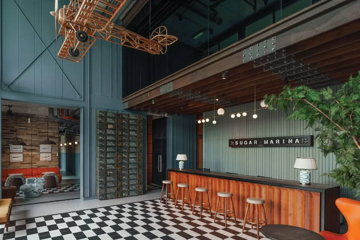 Interior of a modern, stylish bar or cafe with a checkered tile floor, a bar counter with five stools, and walls decorated with wood and vertical paneling. There are two lamps on the bar, a small tree on the right, a wooden ceiling feature above, and a model airplane hanging from the ceiling. The sign on the wall reads 'Sugar Marina.'
