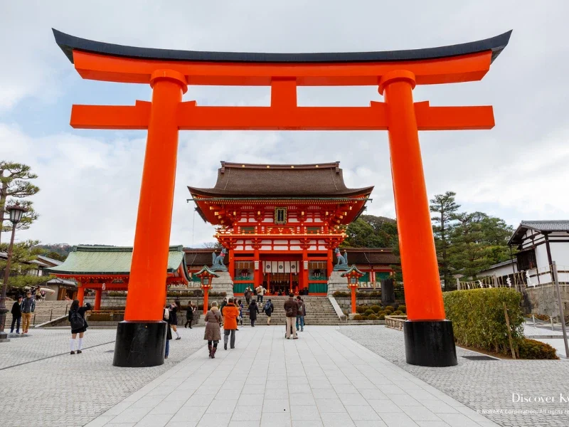Large red torii gate leading to a traditional Japanese shrine with people walking around.