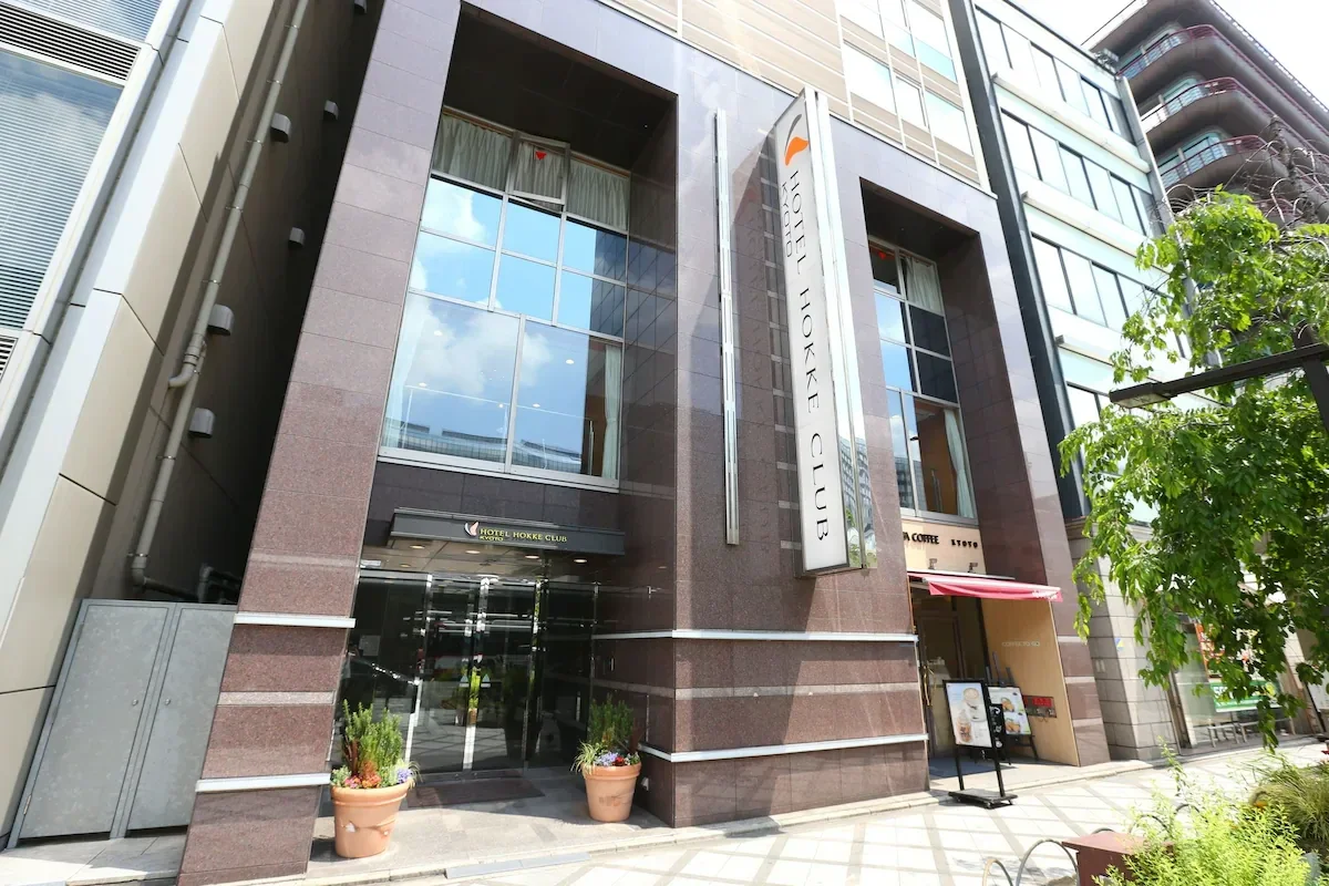 Exterior view of the Hotel Hokke Club with large glass windows, a vertical sign, and potted plants near the entrance in an urban area.