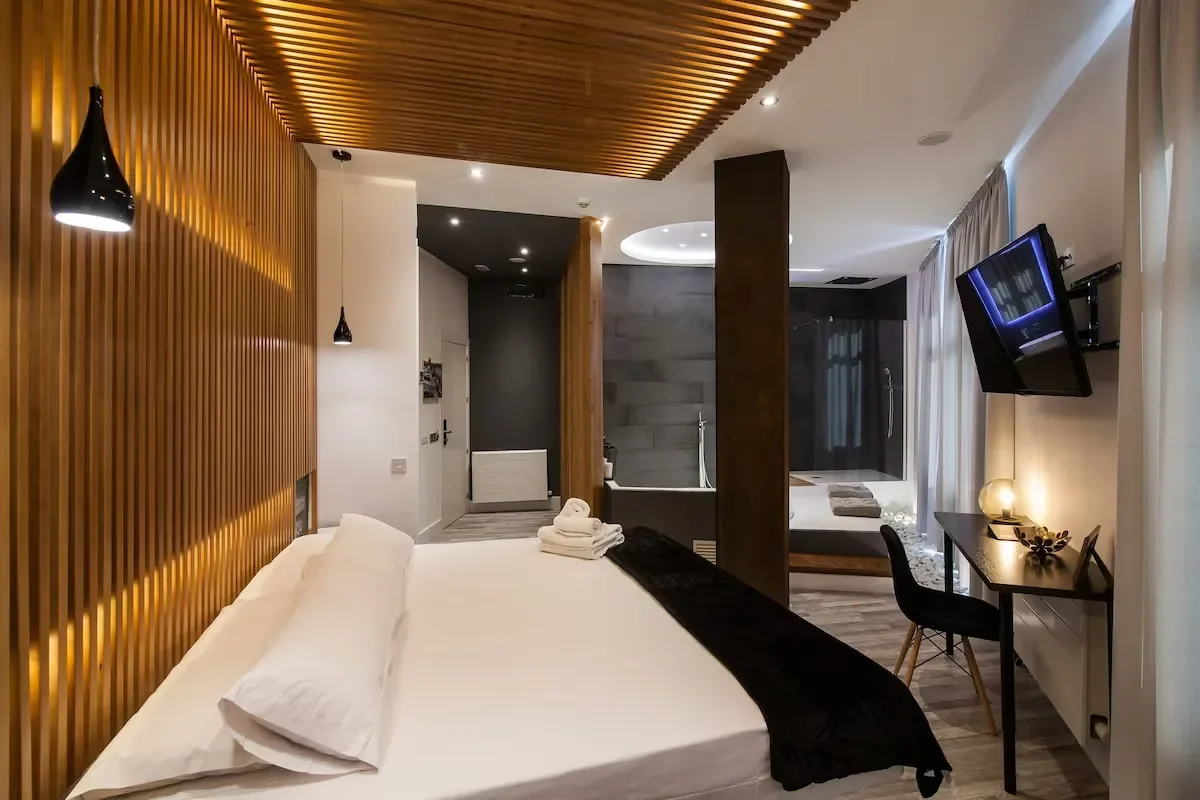 Modern hotel room with a large bed, wooden accent wall, and a bathroom with a bathtub and shower in the background. A flat-screen TV is mounted on the wall, and there is a small desk with a chair and a decorative lamp on it.