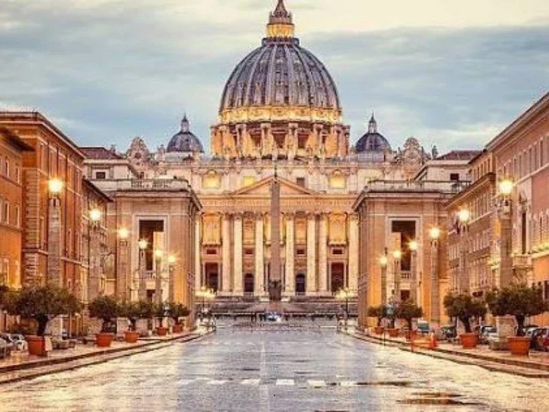 A grand historic building with a large central dome, flanked by symmetrical smaller domes, in a city street setting, illuminated by streetlights.
