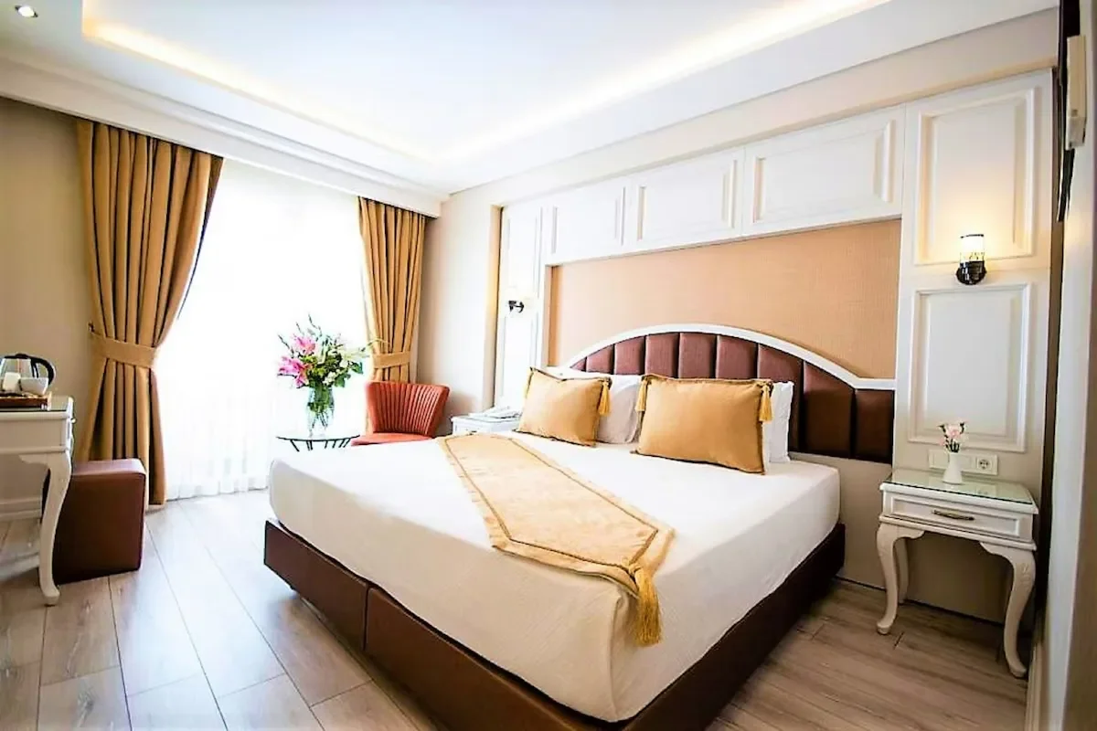 Hotel room with a large bed, two gold pillows, beige walls, a window with heavy curtains, a side table, a chair, and a vase of pink flowers.