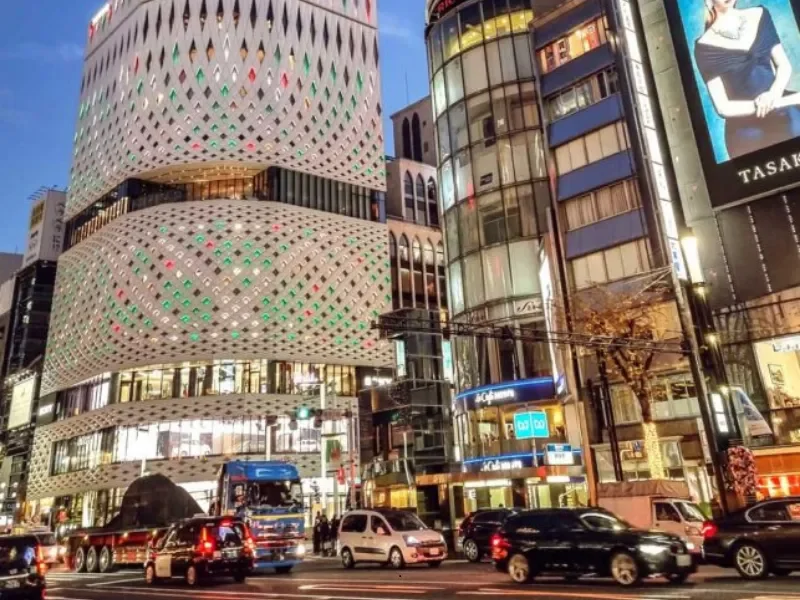 Night city street scene showing modern high-rise buildings with digital advertisements and illuminated signs, with traffic including cars and trucks at the intersection.