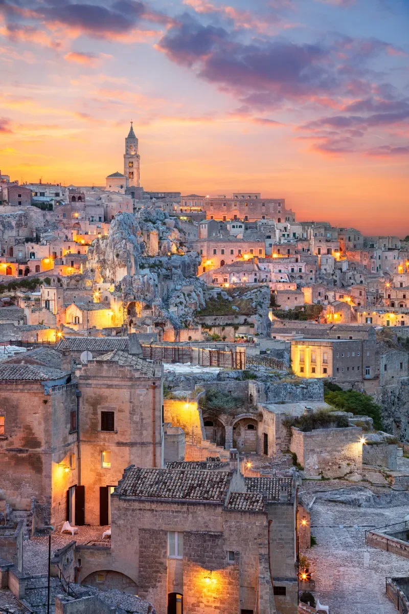 Aerial view of ancient stone buildings in a hillside town during sunset, with warm yellow lights illuminating the narrow streets and a tall clock tower in the background under a colorful sky with pink and purple clouds.
