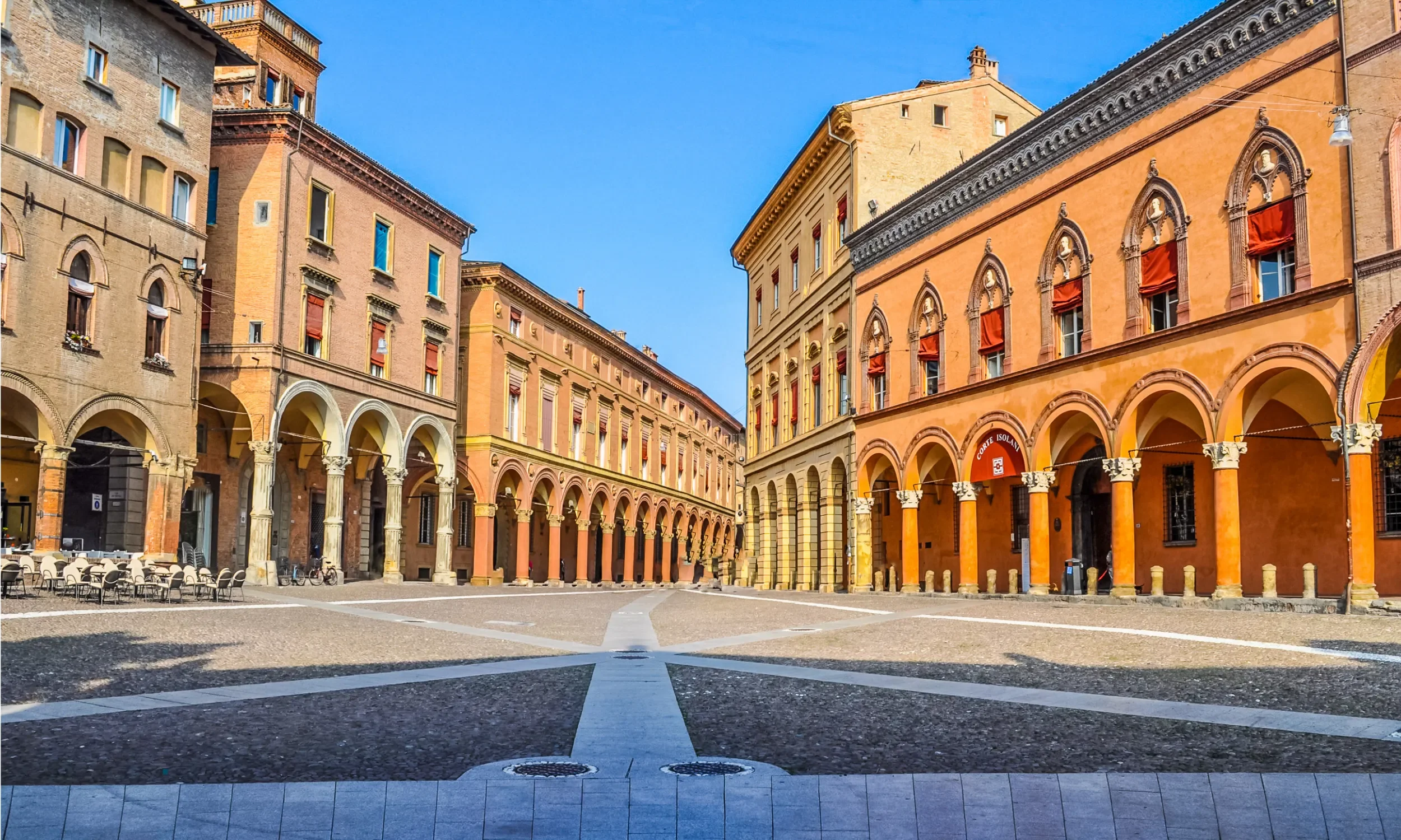 A sunny city square with historical buildings, arched walkways, outdoor café tables, and empty chairs, under a clear blue sky.
