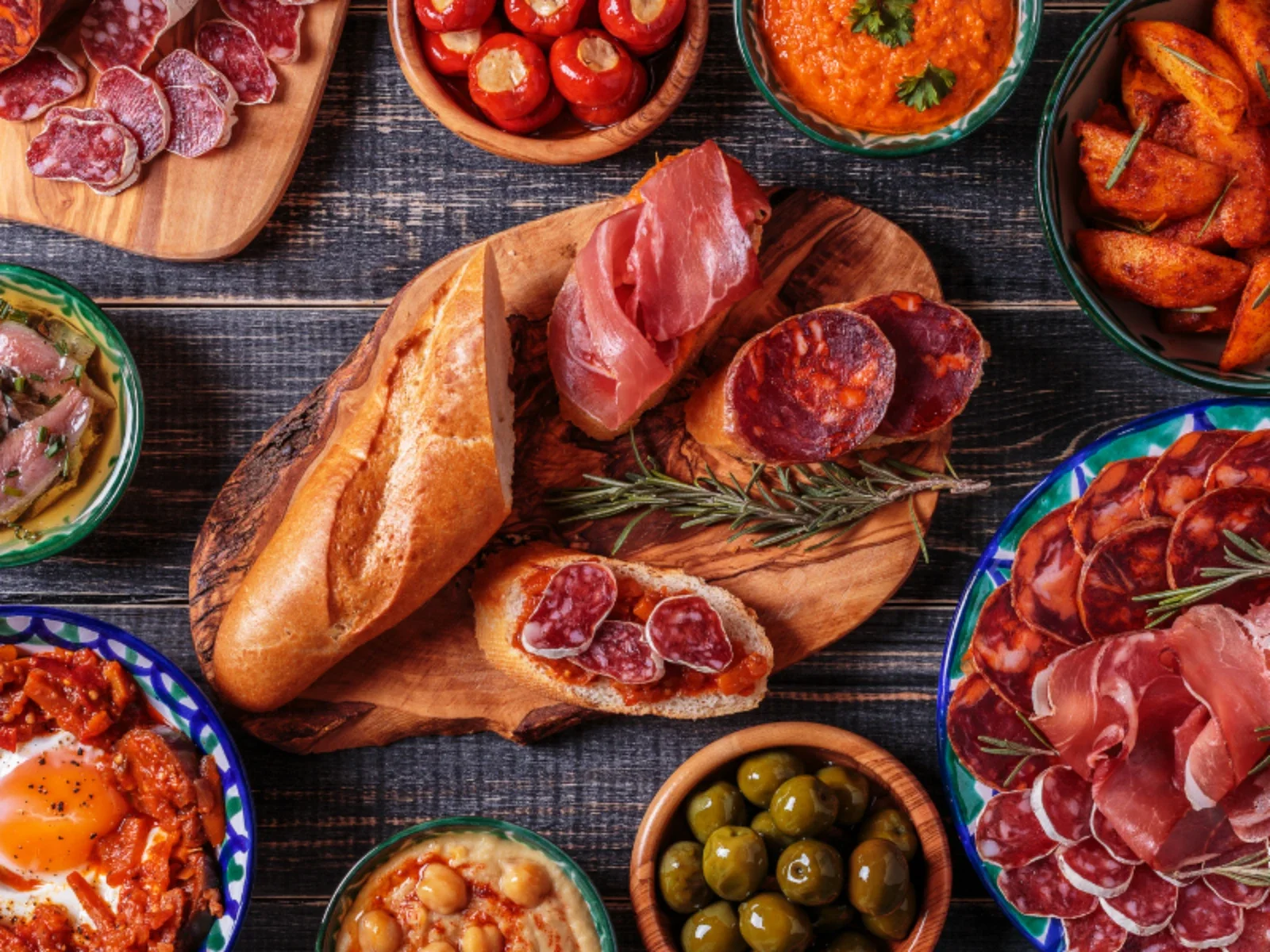 A variety of Mediterranean dishes on a dark wooden table, including bread, sliced cured meats, olives, roasted potatoes, and tomato-based dips and spreads.