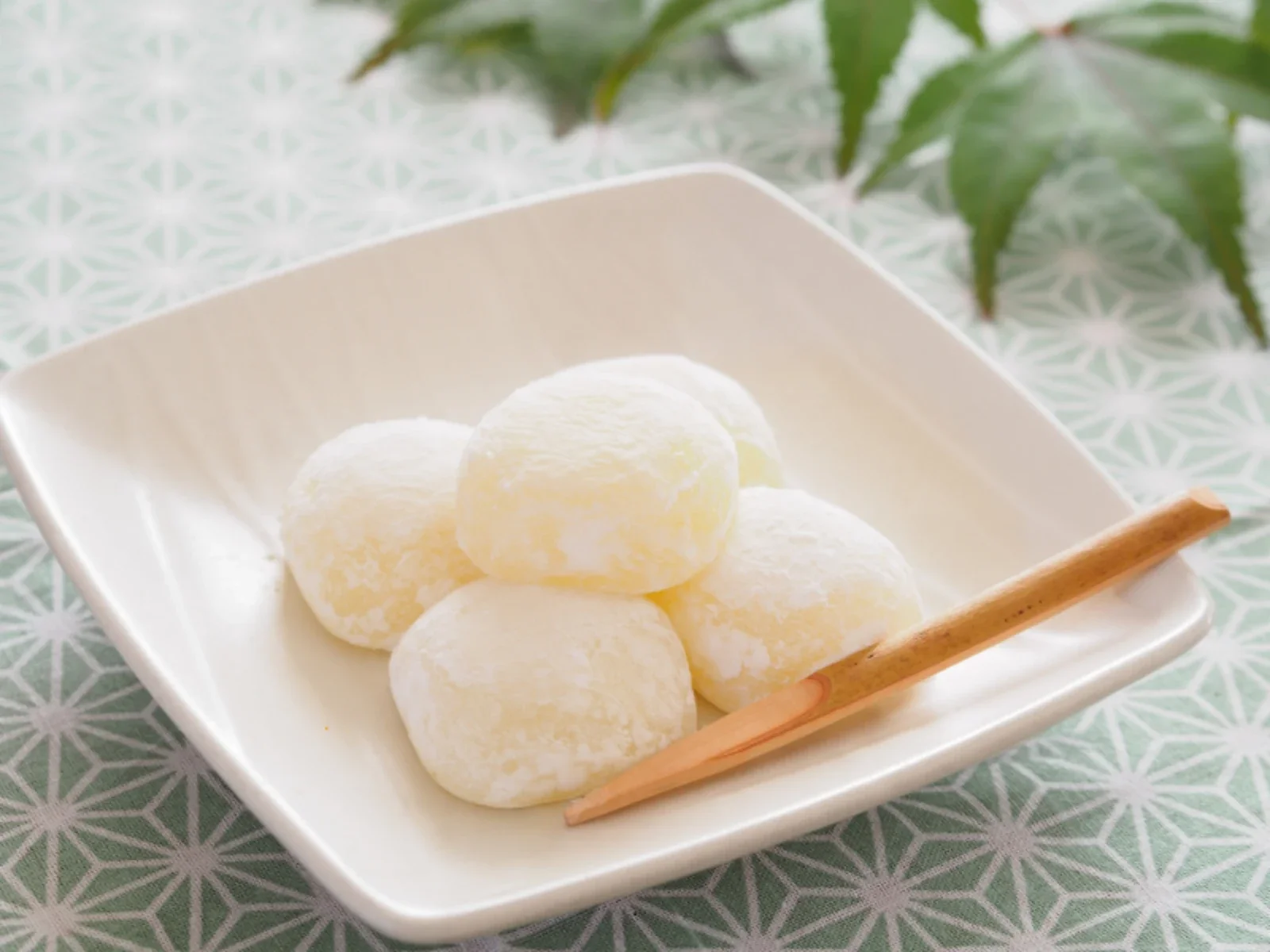 A white square dish containing six white candy balls with a powdered coating, accompanied by a wooden spoon, set on a patterned tablecloth with green leaves in the background.