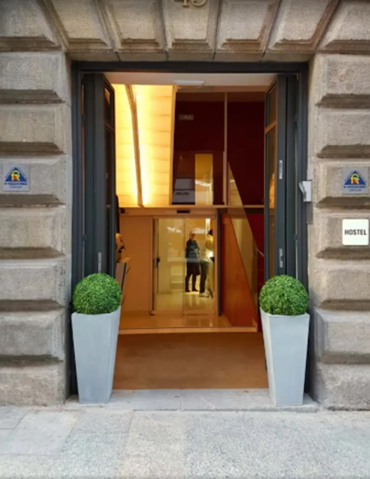 Entrance to a building with large glass doors, flanked by two potted green plants, revealing an interior with warm lighting and a staircase. There are two people reflected in the glass, and a white sign with 'HOSTEL' written on it is on the right side.