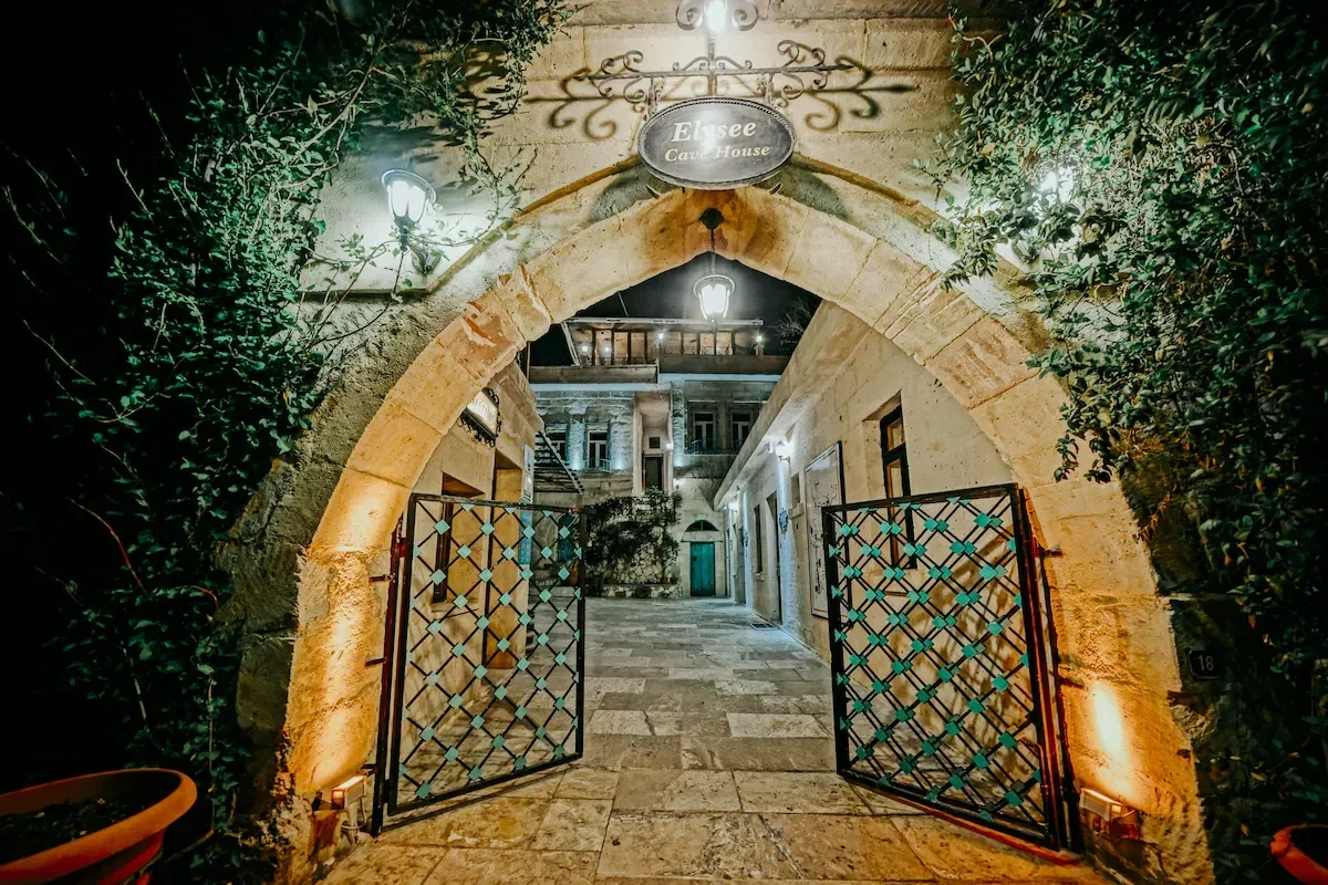 Night view of the entrance to a charming stone building with an archway, illuminated by hanging lanterns, with greenery on the sides and a sign that reads 'Elisee Cave House'.
