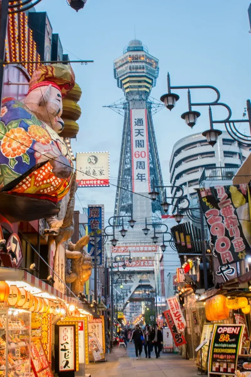 Street scene in Japan with illuminated signs, food stalls, and a tall tower in the background, possibly Osaka. A large decoration of a woman in traditional clothing is on the left.