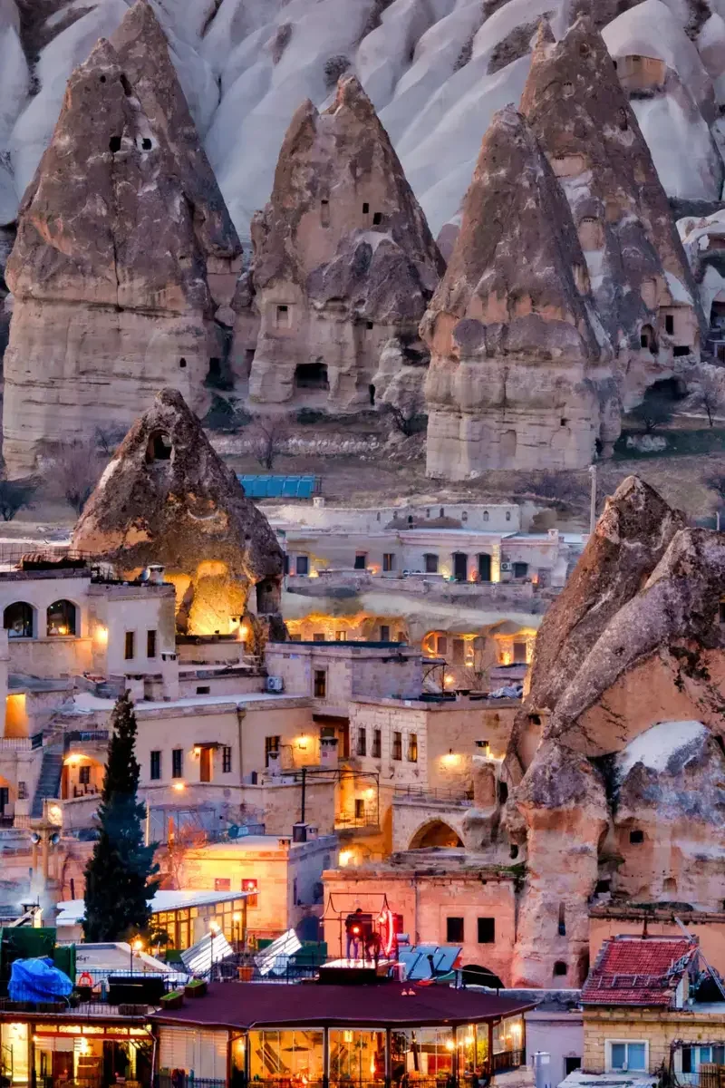 Rock formations with ancient cave dwellings above a town with illuminated buildings at dusk.