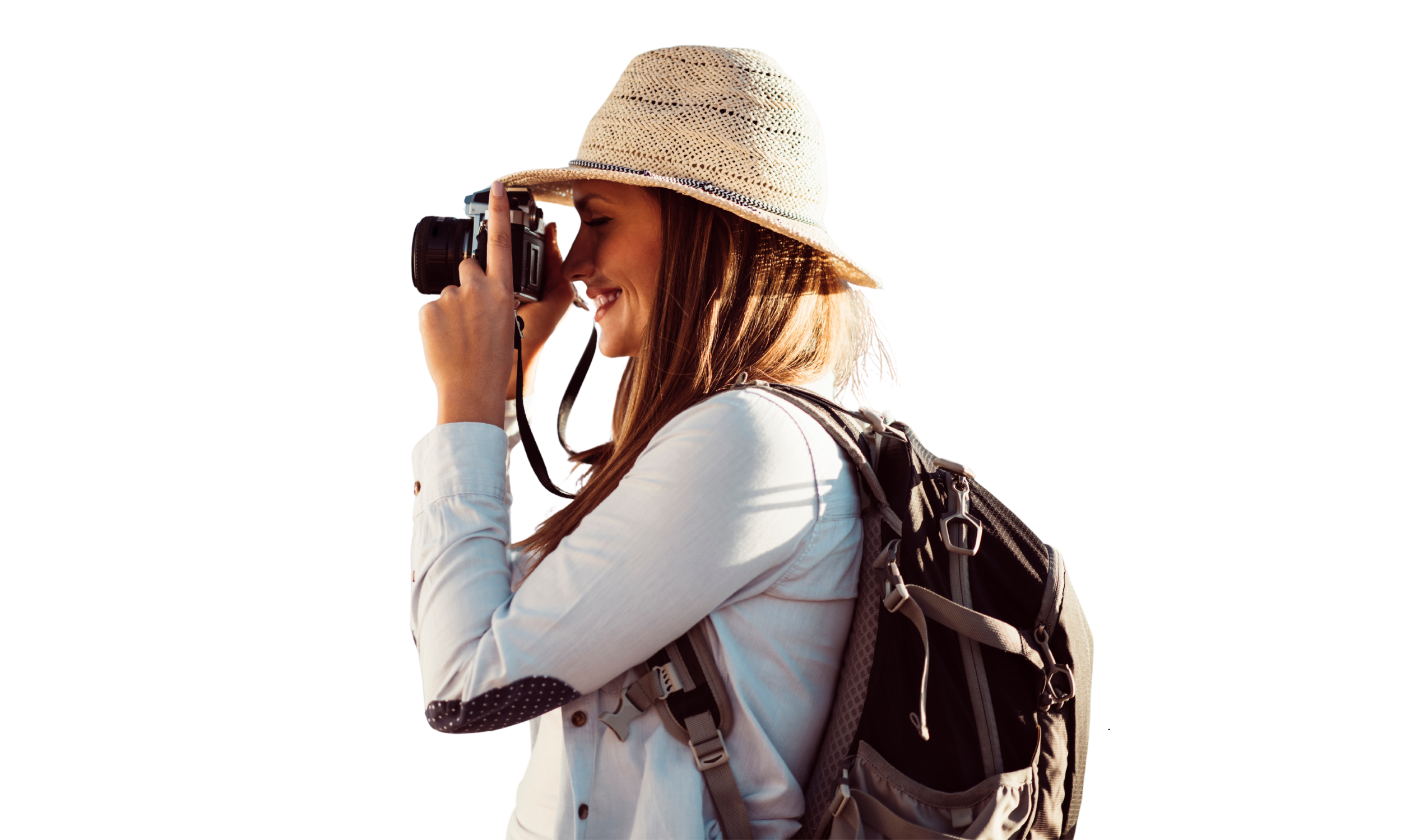 A woman wearing a straw hat and white shirt is taking a photograph with a digital camera. She has a backpack on her back and is standing outdoors, smiling with her eyes closed.
