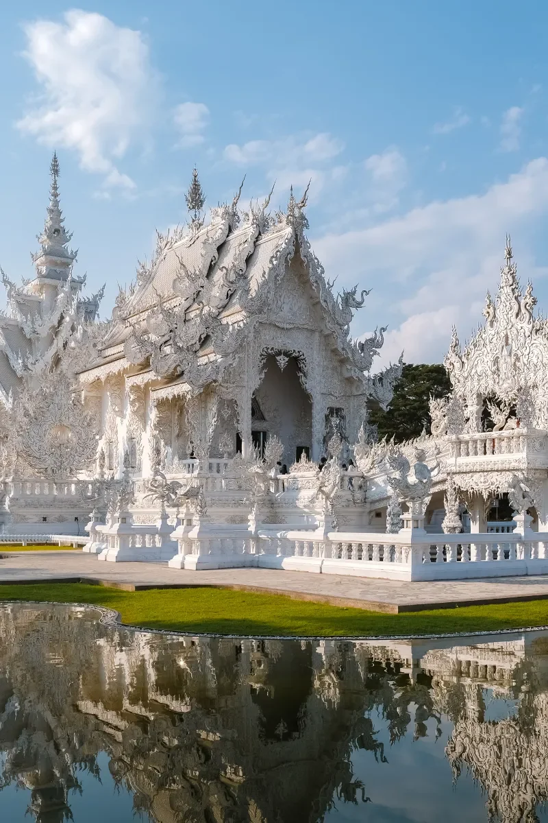 White ornate temple with intricate carvings, surrounded by a small pool reflecting the structure, set against a blue sky with clouds.