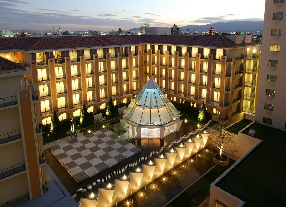 A view of a hotel courtyard at dusk with illuminated windows and a glass pyramid-shaped structure in the center, surrounded by balconies and trees.
