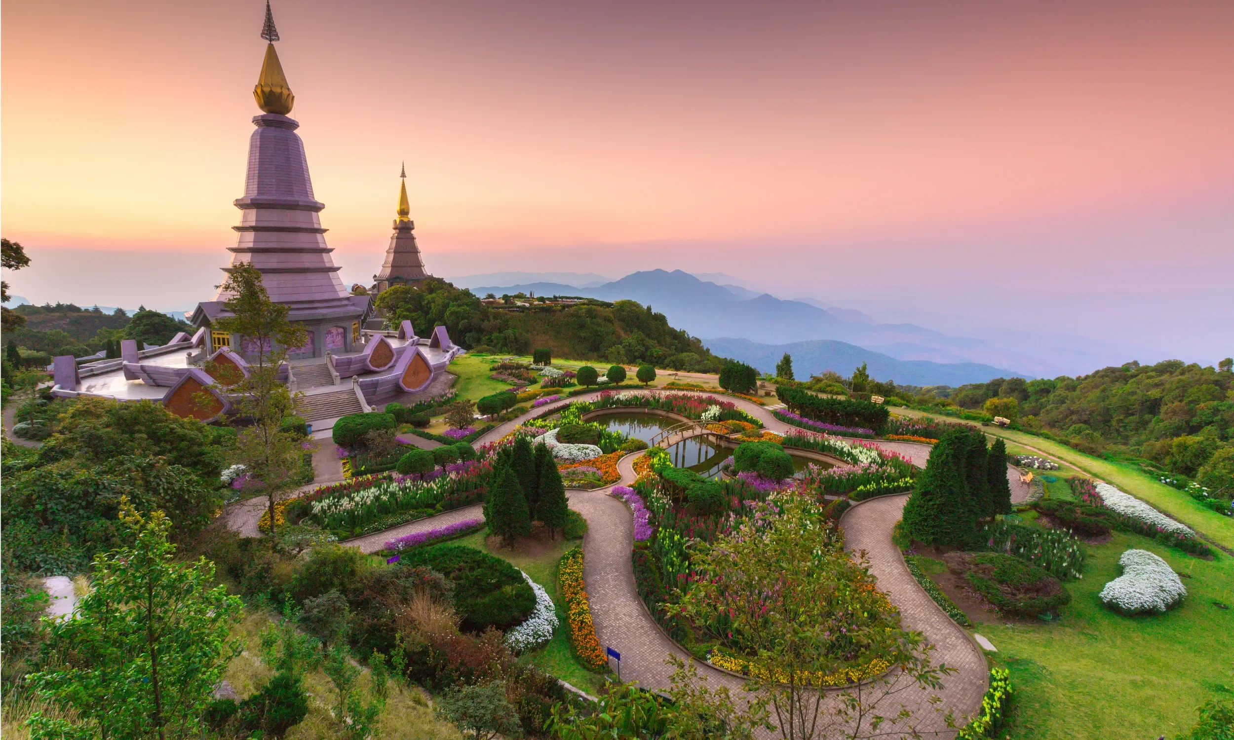 A scenic view of a series of ornate, tiered pagoda-style temples with golden spires, set atop a lush green hill. The foreground features a vibrant, well-maintained flower garden with a winding pathway, small pond, and neatly trimmed bushes. Misty mountains are visible in the distance under a pastel-colored sky during sunset or dawn.