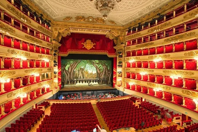 View of a grand theater with ornate gold and red decor, a painted stage with a forest scene, and multiple balconies filled with red velvet seating.