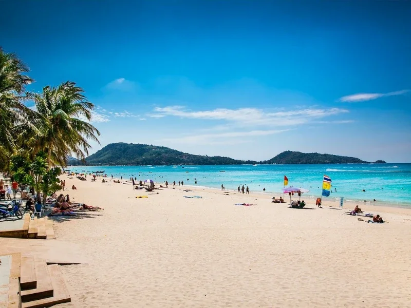 Sunny beach with palm trees, people sunbathing and swimming, colorful umbrellas, and a distant island or landmass in the background under a blue sky.
