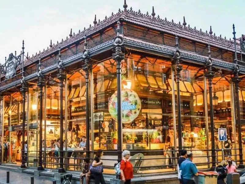 A historic glass and iron building with ornate black metalwork, illuminated from within, with people walking outside.