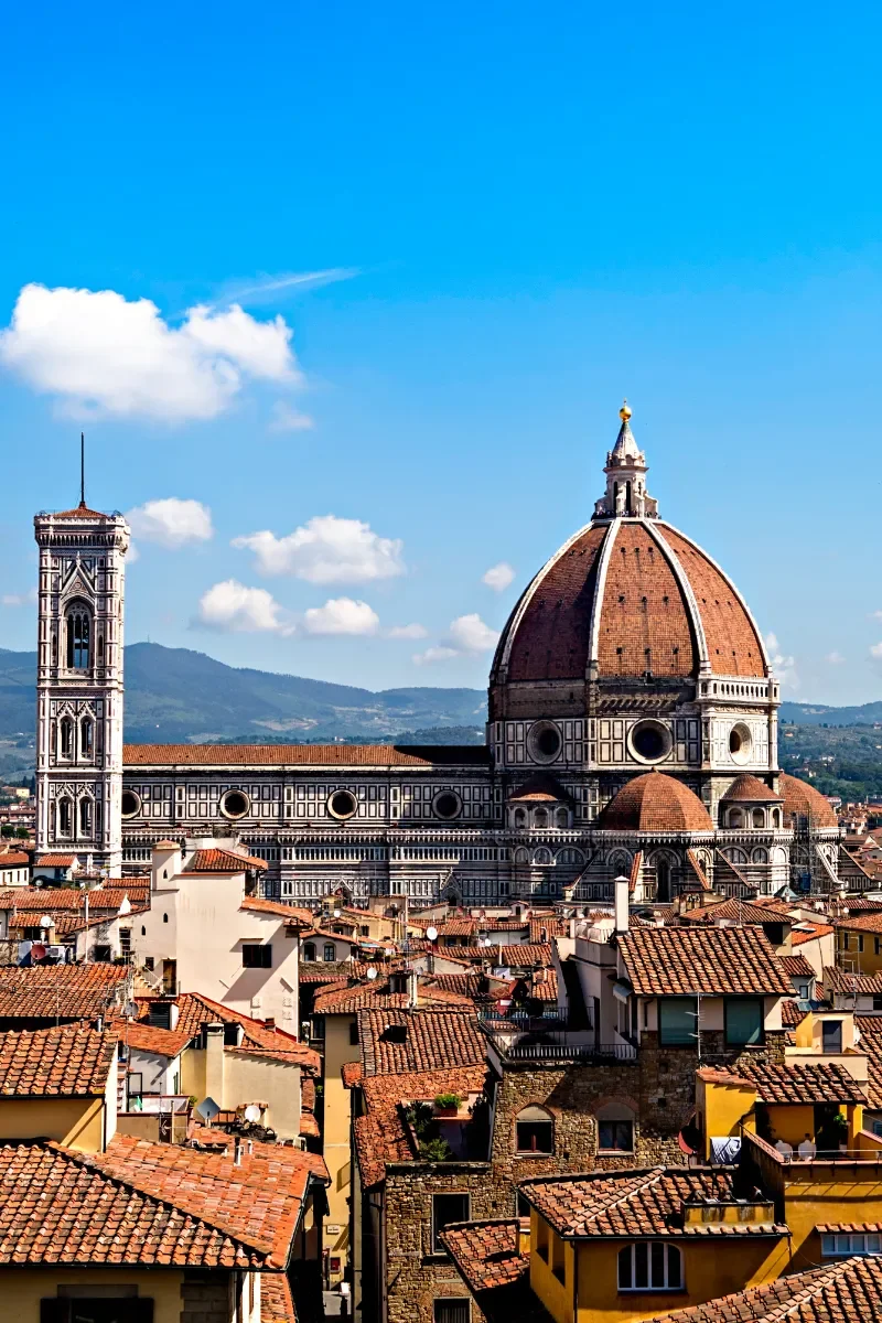 View of Florence, Italy, with the Florence Cathedral's dome and bell tower visible against a blue sky with clouds, surrounded by red-tiled rooftops.