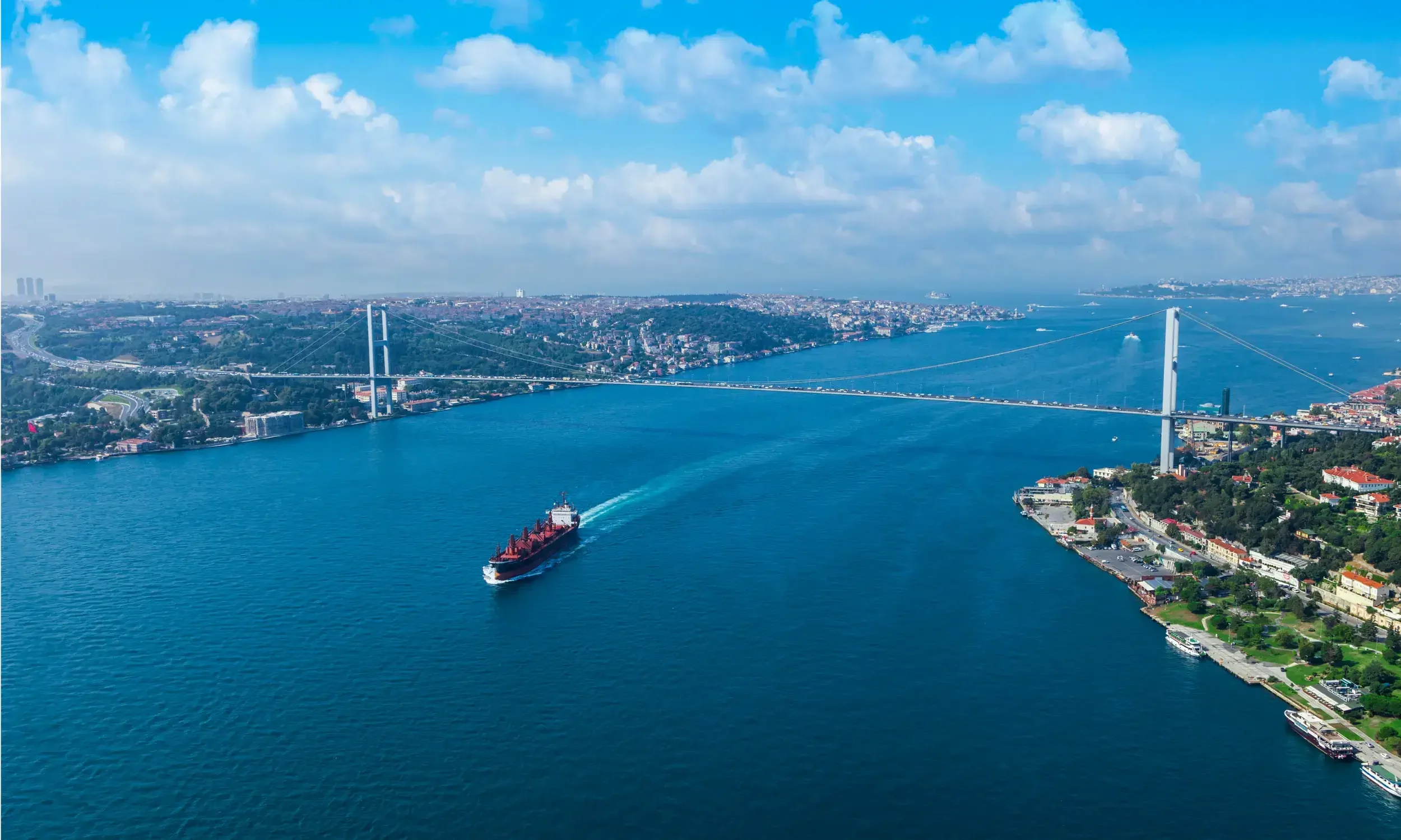 Bosphorus Aerial view of a large body of water with a cargo ship sailing and a bridge connecting city areas on both sides, with a cityscape visible in the background under a partly cloudy sky.
