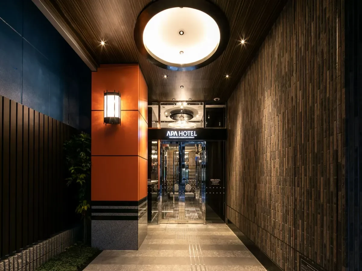 Entrance to APA Hotel with glass doors, illuminated sign, and modern lighting fixtures in the hallway.