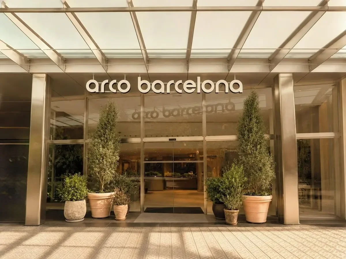 The entrance of arco barcelona is shown with glass doors and tall potted plants on either side.