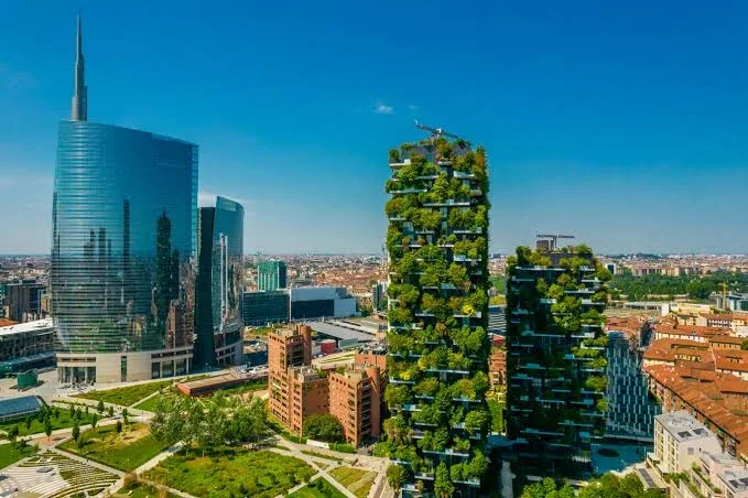 Cityscape with modern skyscrapers, including a large glass building and a unique green rooftop building with terraces filled with trees and plants under a clear blue sky.