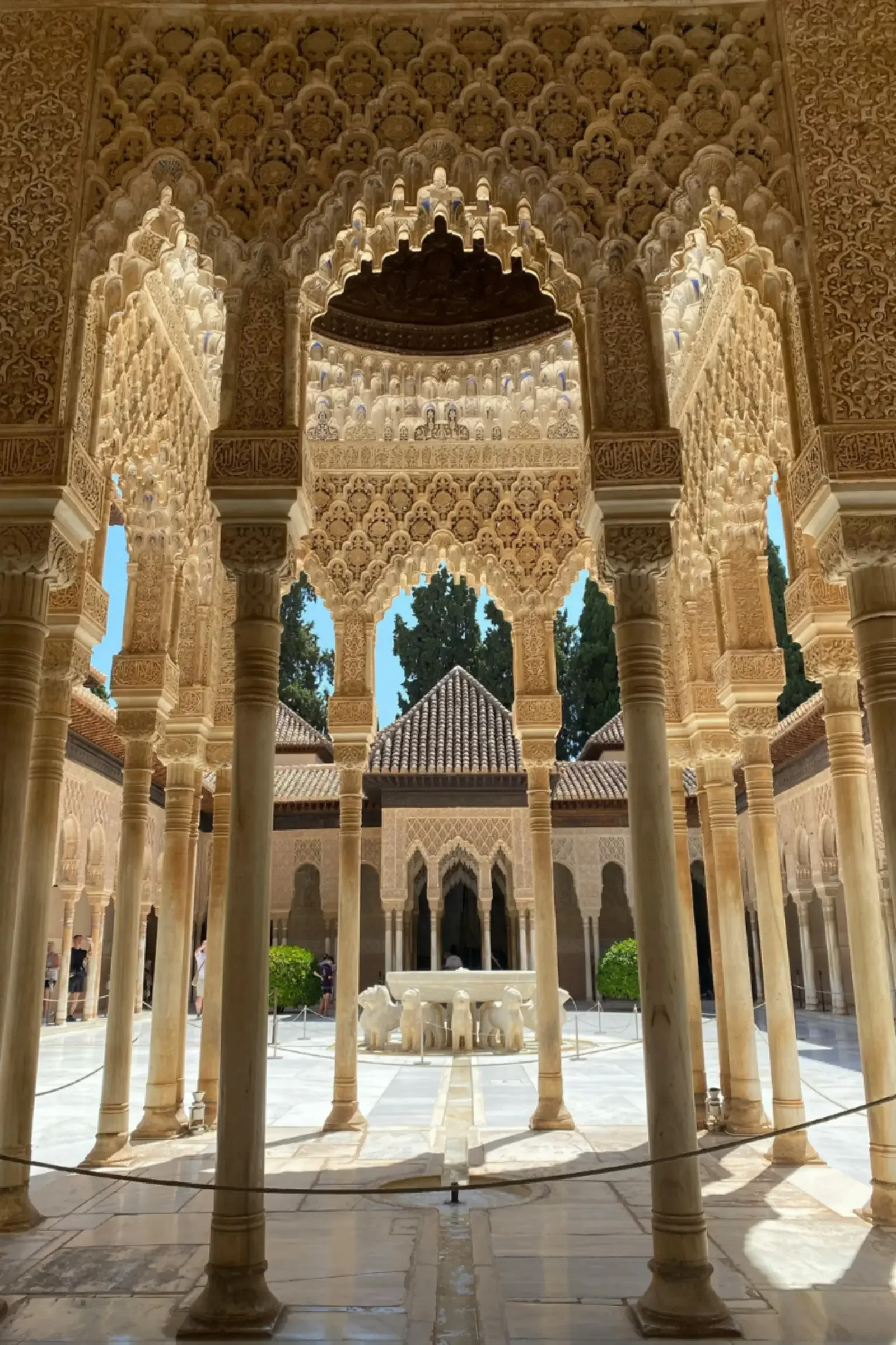 Courtyard with white marble fountain, surrounded by intricate carved stone columns and walls, with a view of lush trees and traditional tile roof structures in the background, reflecting Islamic architecture.