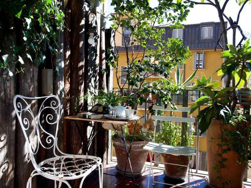 Small balcony with potted plants, a white metal chair, and a wooden table with tea set, overlooking neighboring yellow building.