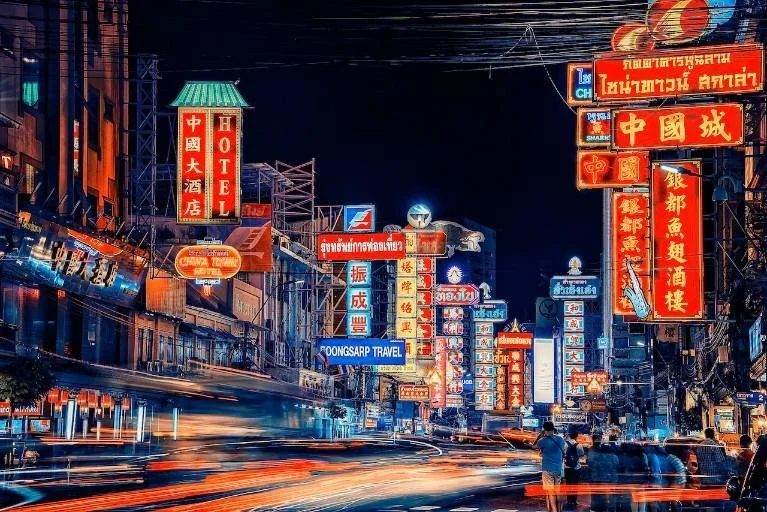 Nighttime street scene with illuminated signs in multiple languages, including Chinese and Thai, and blurred moving cars.