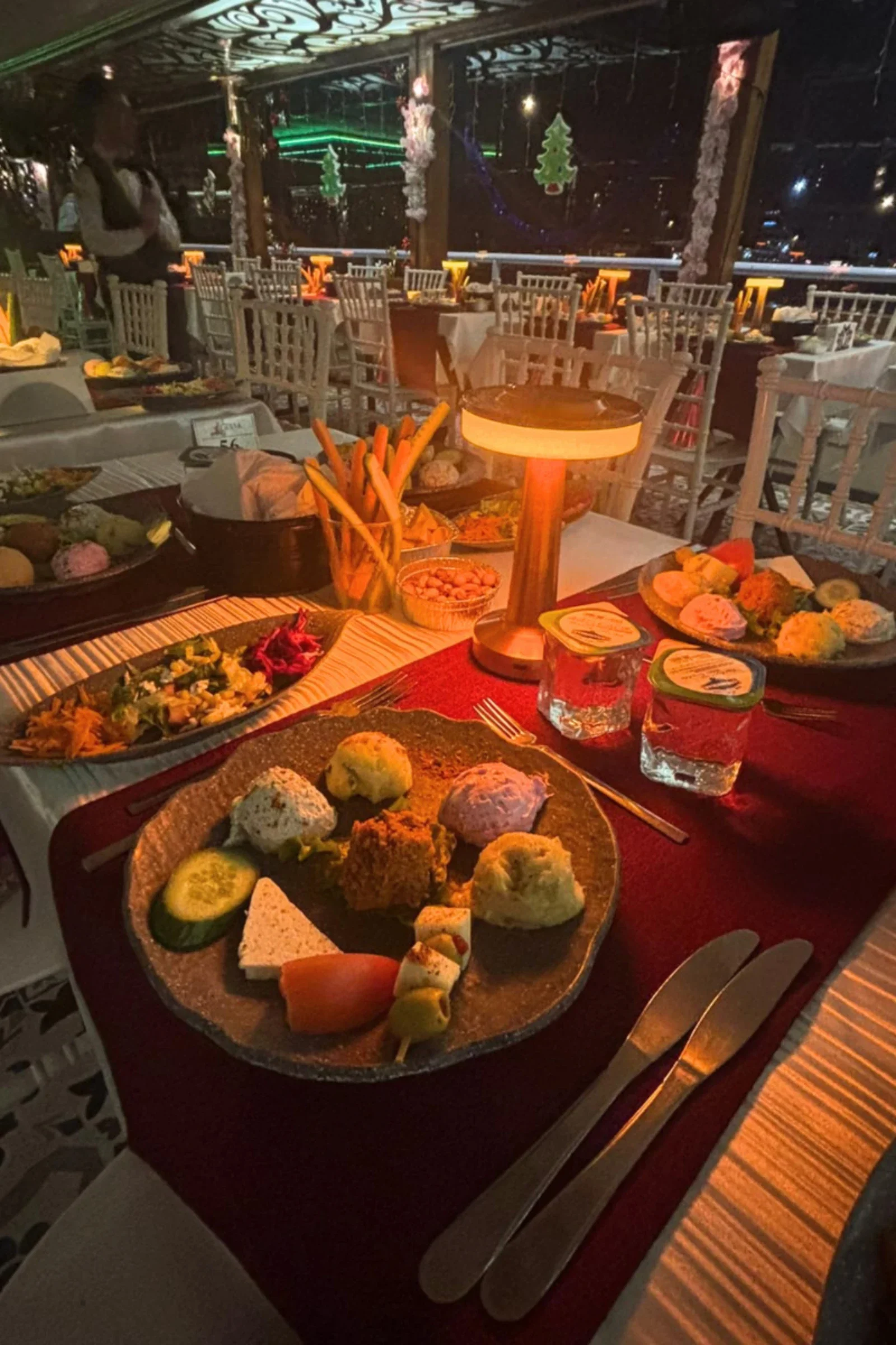 A restaurant table set with various dishes, including bread rolls, vegetables, and salads, on a red tablecloth with a small wooden lamp, in a decorated dining area with Christmas ornaments and chairs.