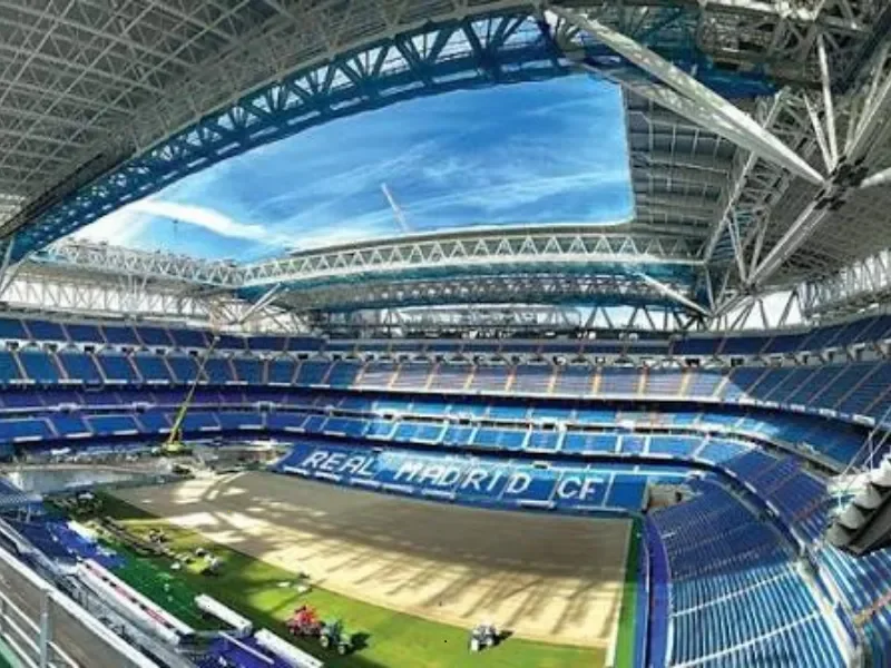 Inside a large, modern soccer stadium with blue seating and a partially open roof, showing the field and seating area with "Real Madrid CF" on seats.