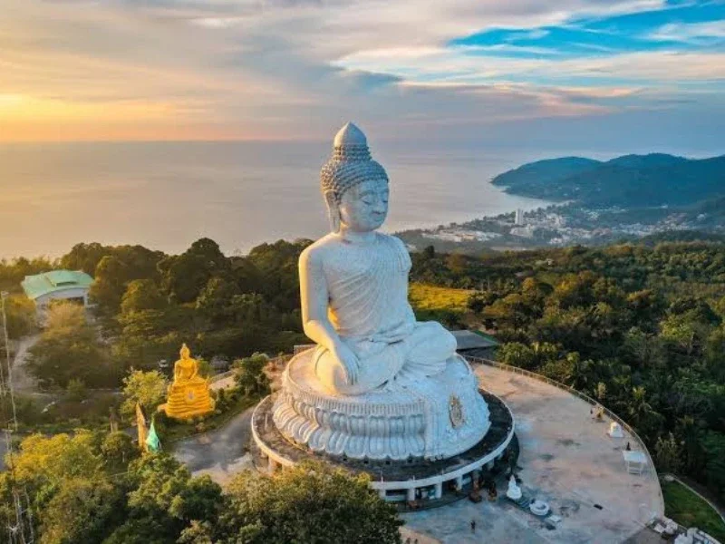 Large white Buddha statue in a seated meditation pose on a hilltop overlooking the ocean, with a smaller yellow Buddha statue nearby and lush green trees surrounding the area.
