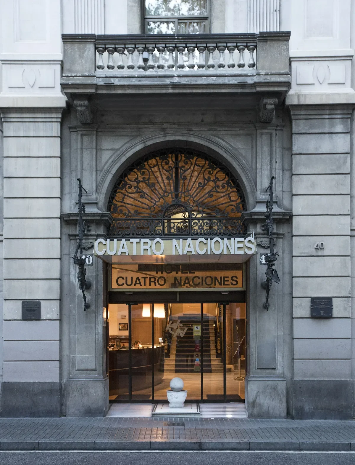The entrance of a hotel named 'Cuatro Naciones', featuring an ornate iron gate, a stone facade, and a sign with the hotel name above the doorway. The building has classical architectural details, including balustrades and decorative sculptures.
