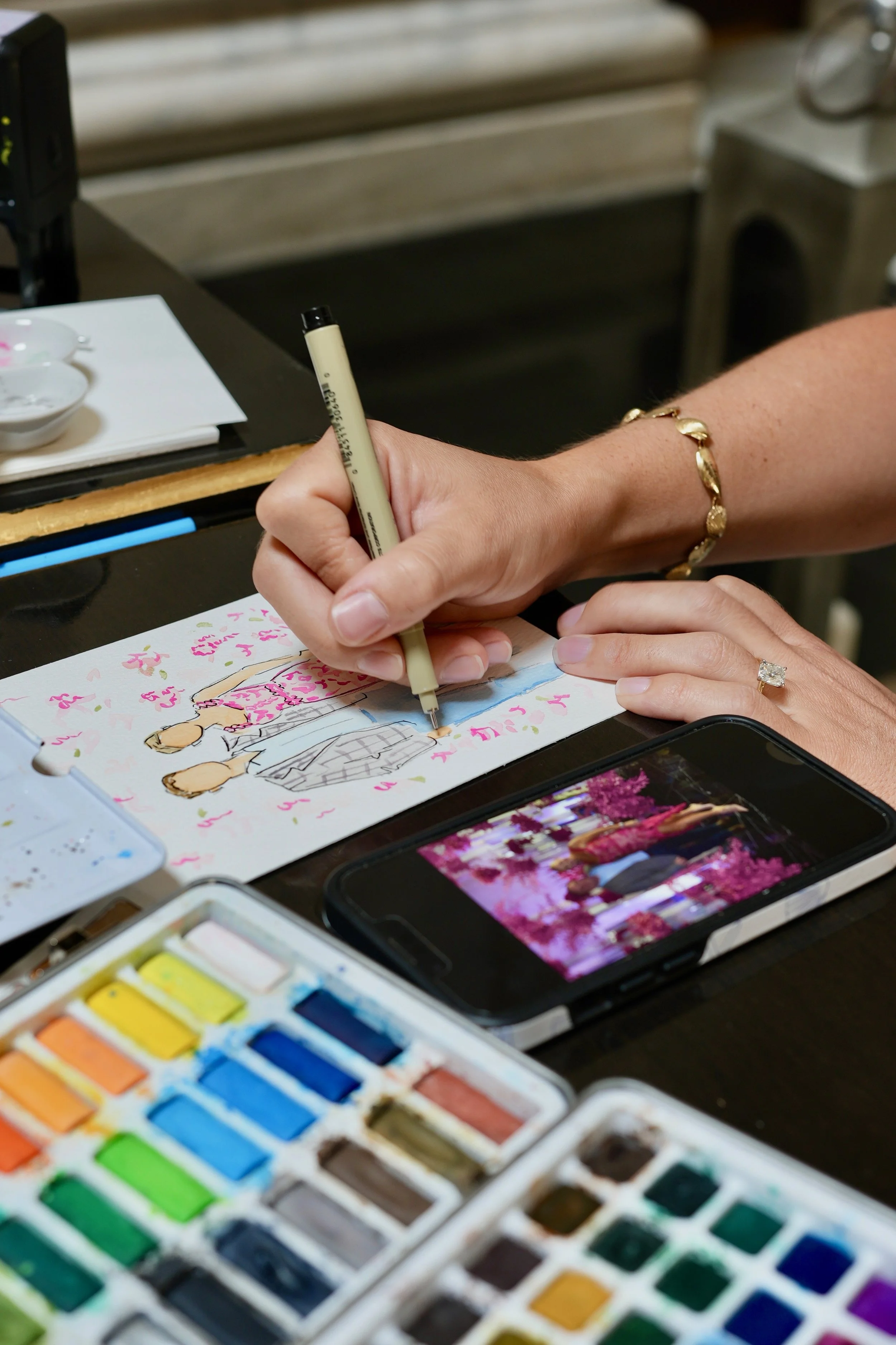Person drawing a fashion sketch with watercolor paints nearby, using a smartphone to view a real-life scene of flowers and people in pink and purple, with art supplies on the table.