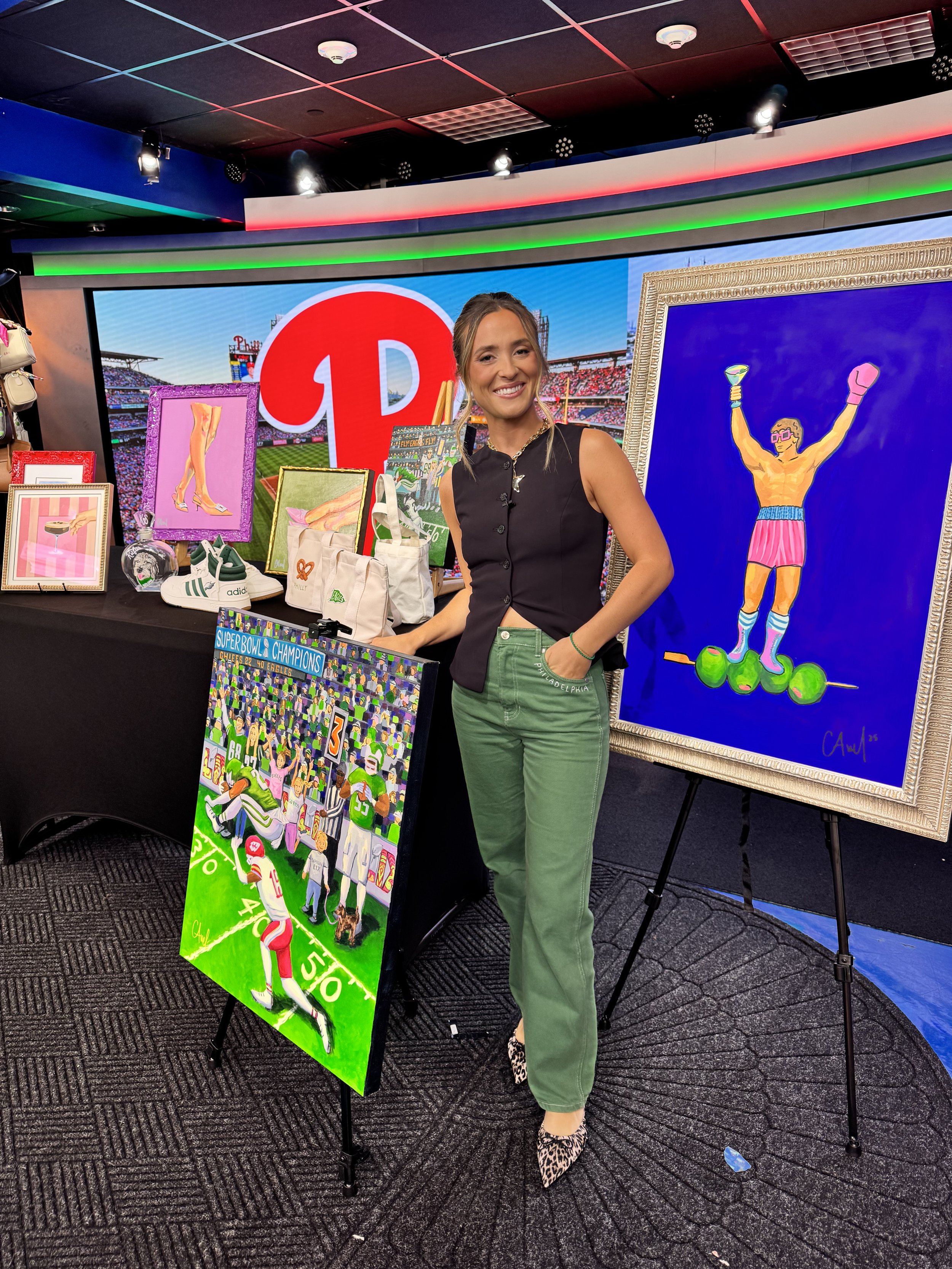 A woman wearing green jeans featuring "Tush Push" on the back pocket. She is smiling, surrounded by her colorful framed artwork including Philadelphias icon Rocky Balboa and a Philadelphia Eagles Super Bowl Champions painting.
