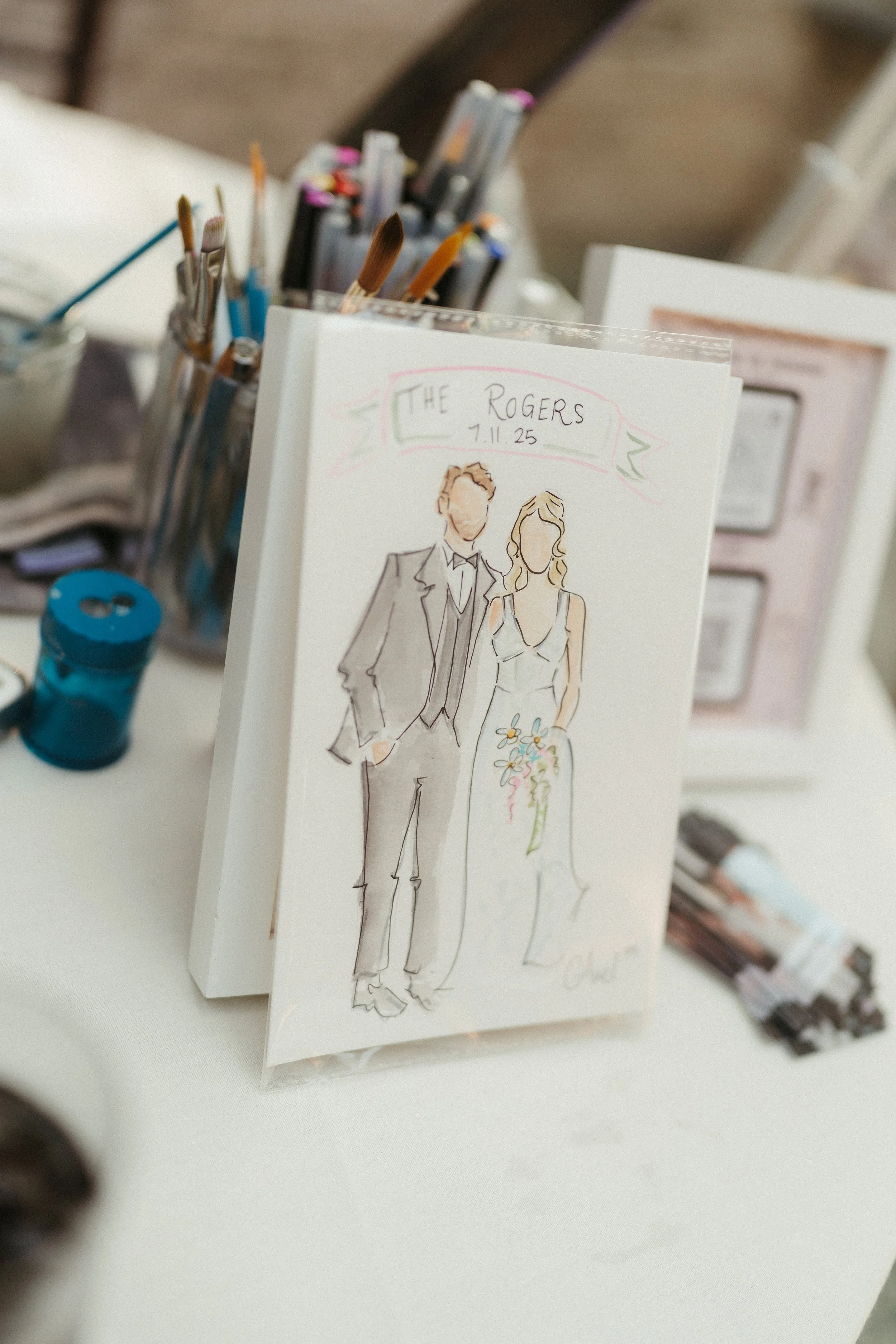 A wedding card featuring a sketch of a bride and groom with the text 'The Rogers, 7.11.25.' surrounded by art supplies on a table. wedding guest portraits. live guest portraits. watercolor portrait.