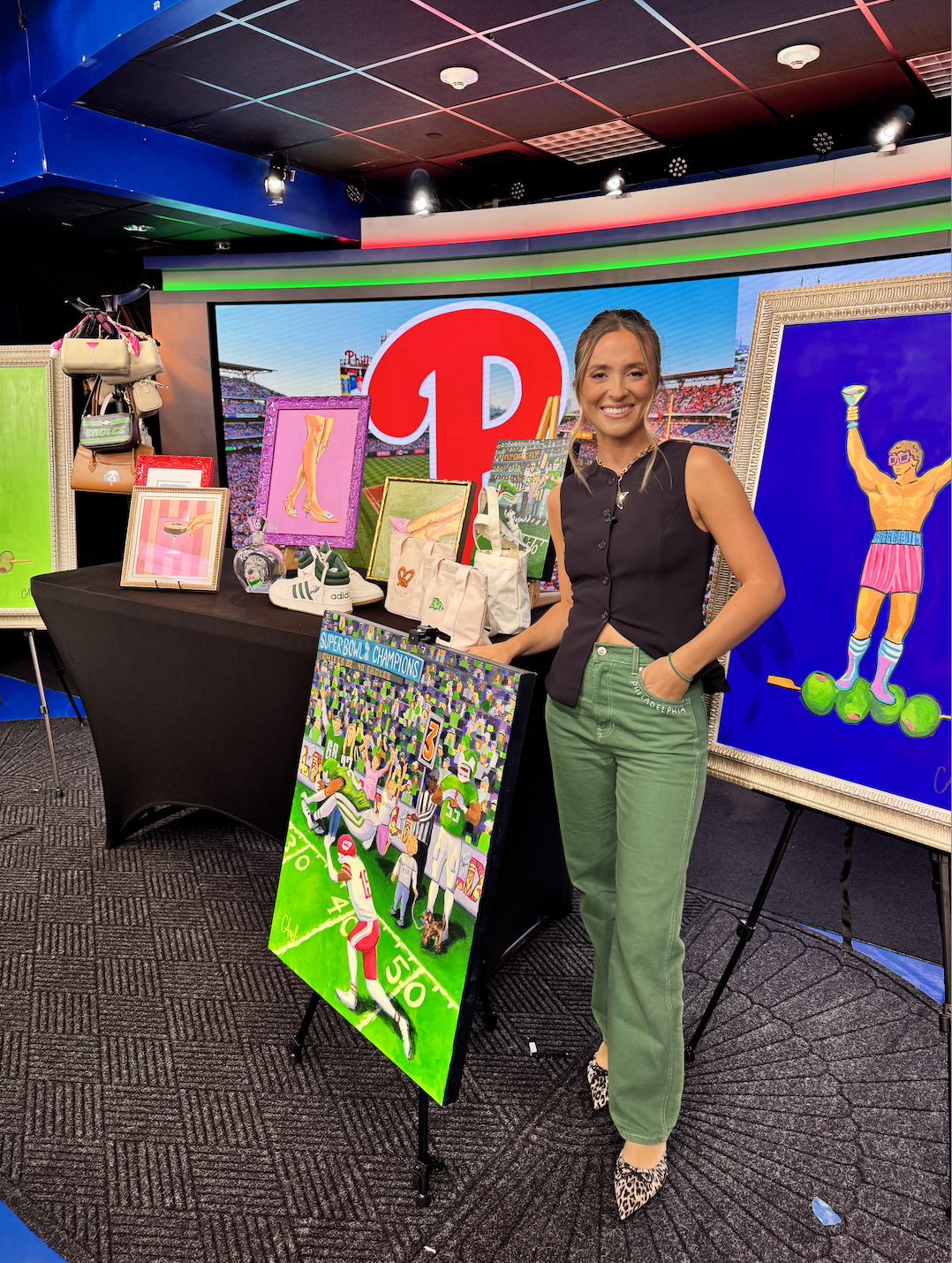 A woman standing next to a display table with various framed art pieces, handbags, and sneakers at an indoor event. There is a large digital screen in the background showing a red logo, and an artwork of a person holding a trophy. The woman is smiling and wearing a black sleeveless vest, green pants, and leopard-print shoes.