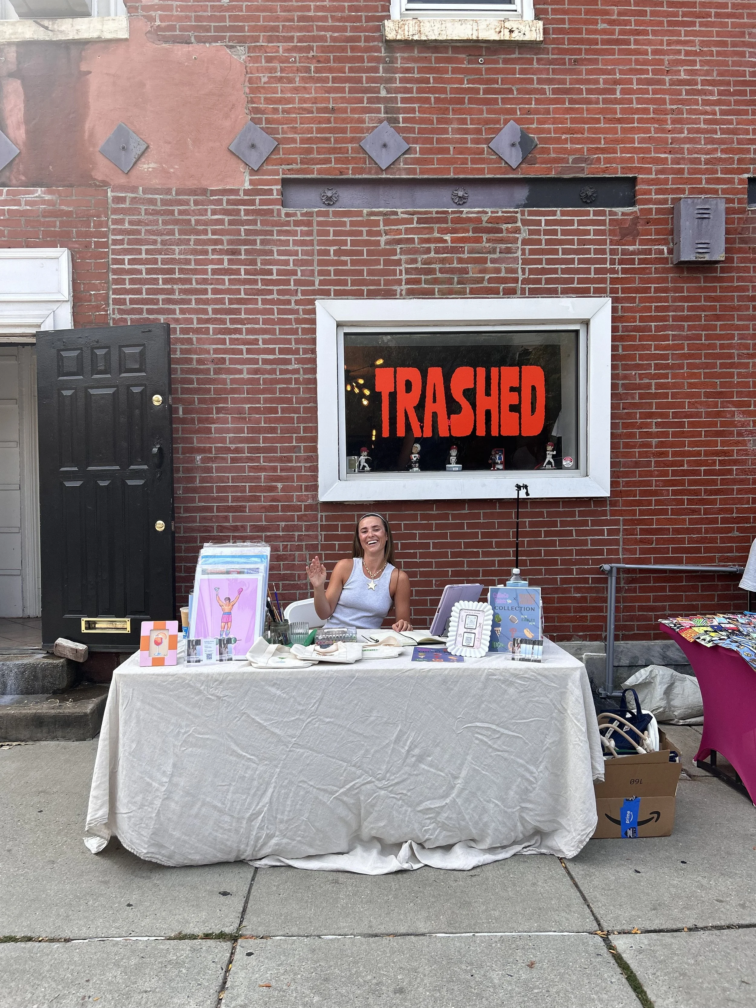Callie is sitting behind a table smiling and waving outside 'TRASHED VINTAGE' boutique. This event is a pop-up selling her artwork and live painting tote bags and other items in Fishtown, Philadelphia, PA.