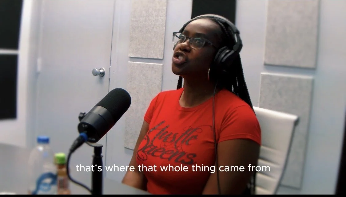 A woman in a red t-shirt speaking into a microphone in a recording studio, wearing large glasses and headphones.