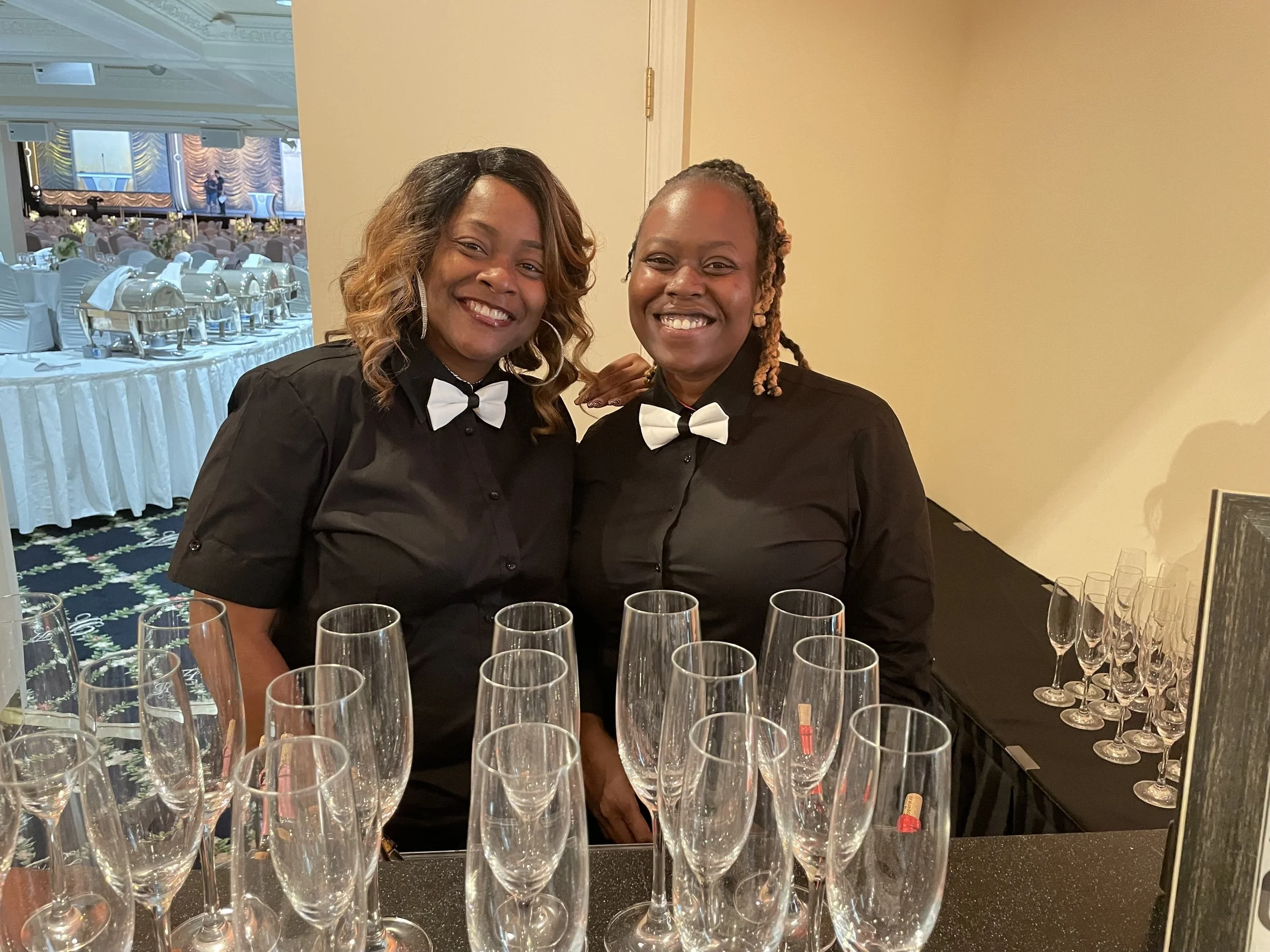 Two women dressed in black shirts with white bow ties, smiling at a beverage station with empty champagne glasses, at a formal event or celebration.