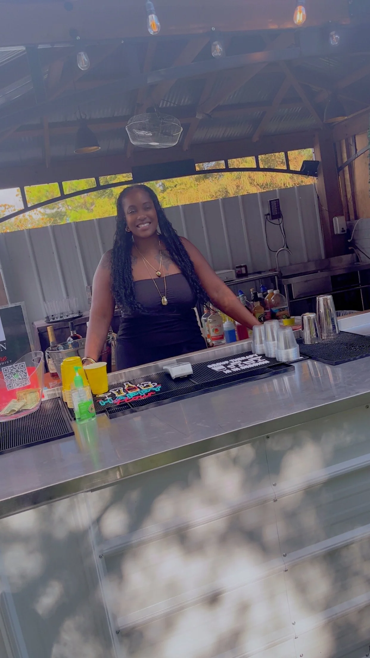A woman with long black hair, wearing a black strapless dress and gold jewelry, stands behind a stainless steel bar counter with various bottles and cups inside an open-air structure with a metal roof.