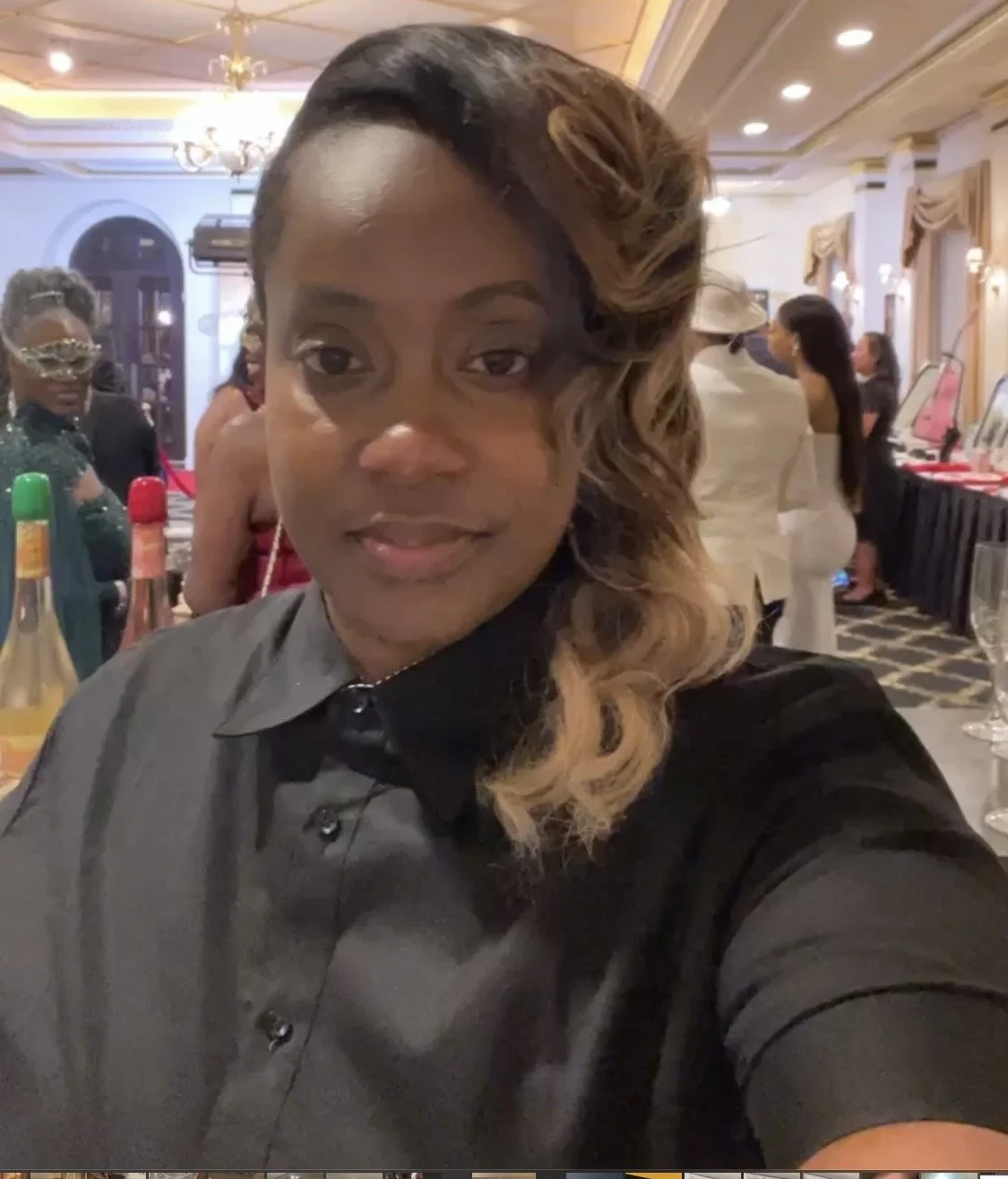 A woman takes a selfie at a social event in an elegant room, with other attendees, bottles, and tables visible in the background.