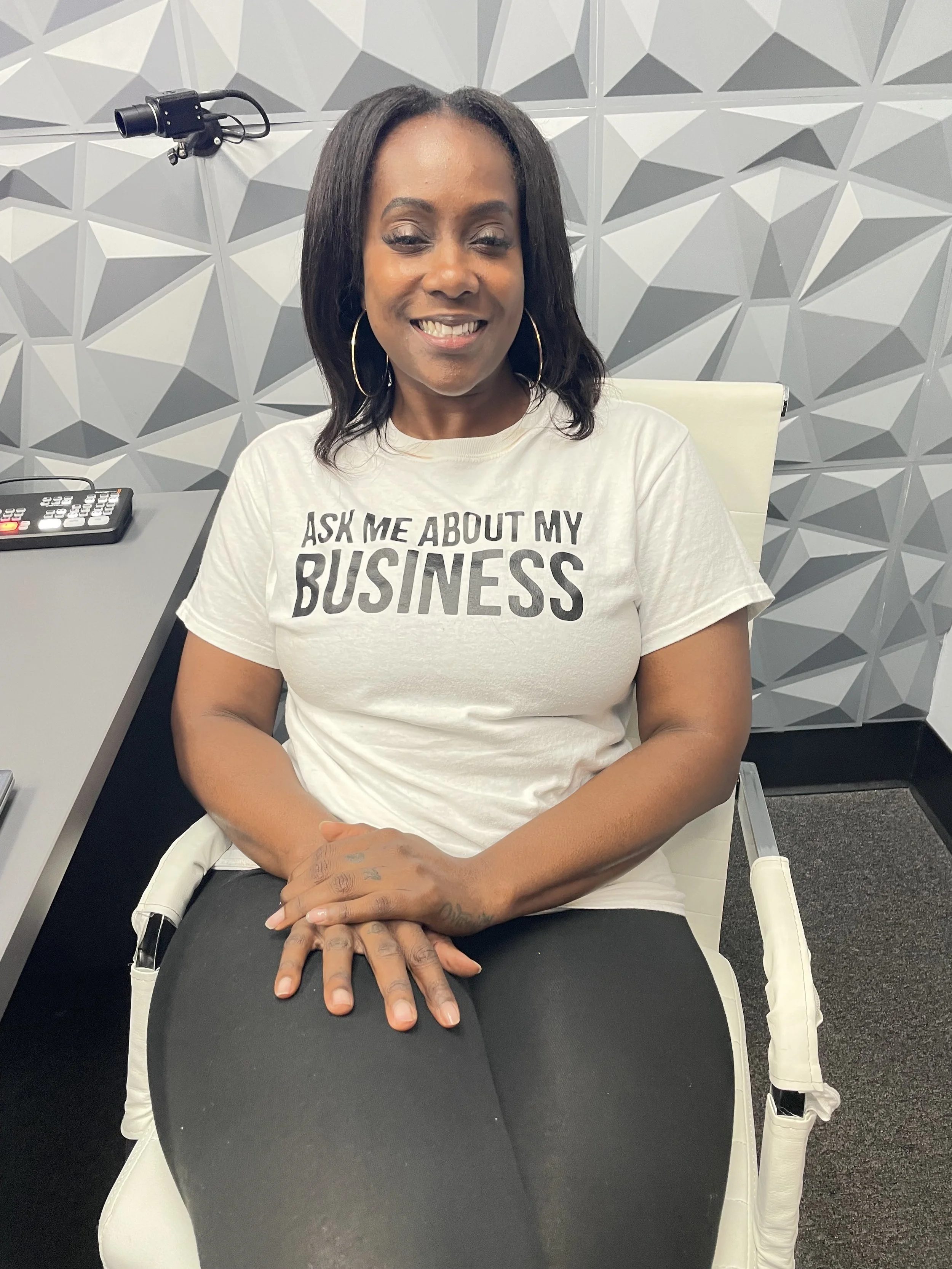 A smiling woman with dark hair, wearing hoop earrings and a white T-shirt with the text 'ASK ME ABOUT MY BUSINESS,' sitting on a white chair in a modern, geometric-patterned gray wall room.