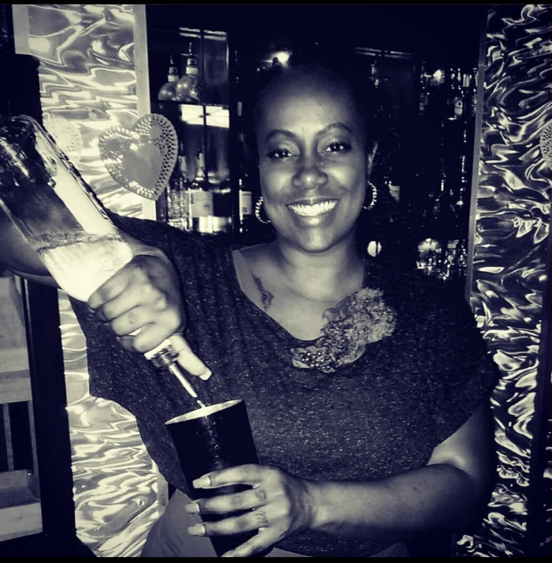 A woman with a big smile pouring a drink from a bottle into a glass at a bar or party.