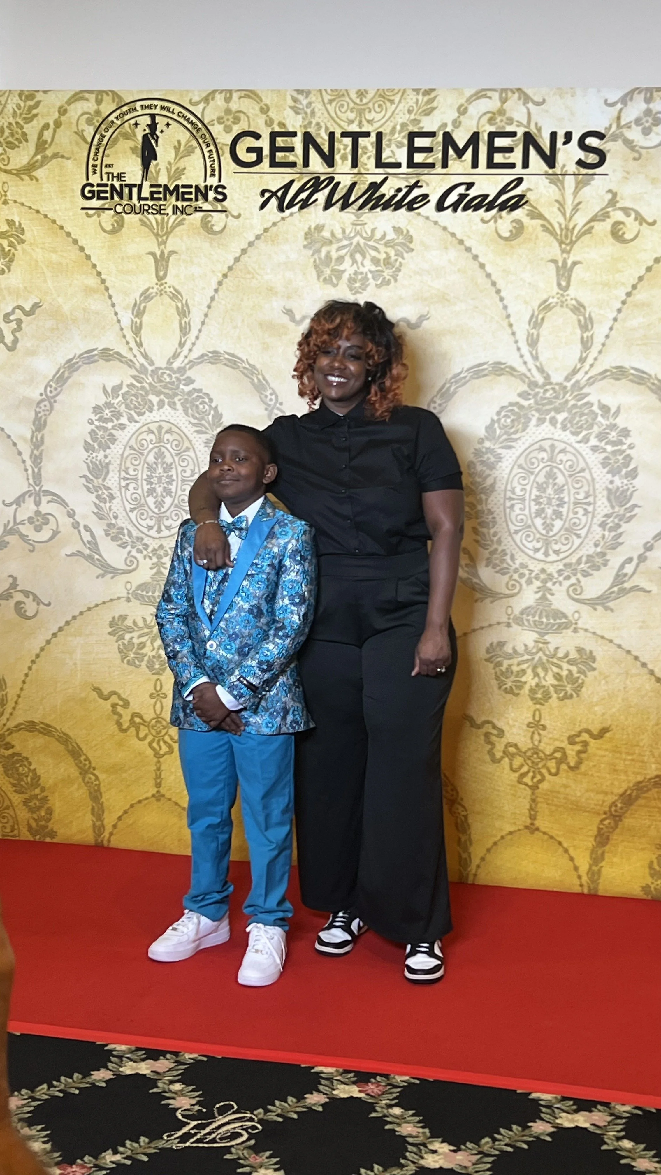 A woman and a young boy standing on a red carpet in front of a backdrop with the text 'Gentlemen's All White Gala' and a logo. The boy is wearing a blue patterned suit with a bow tie, and the woman is dressed in black with black and white sneakers. T