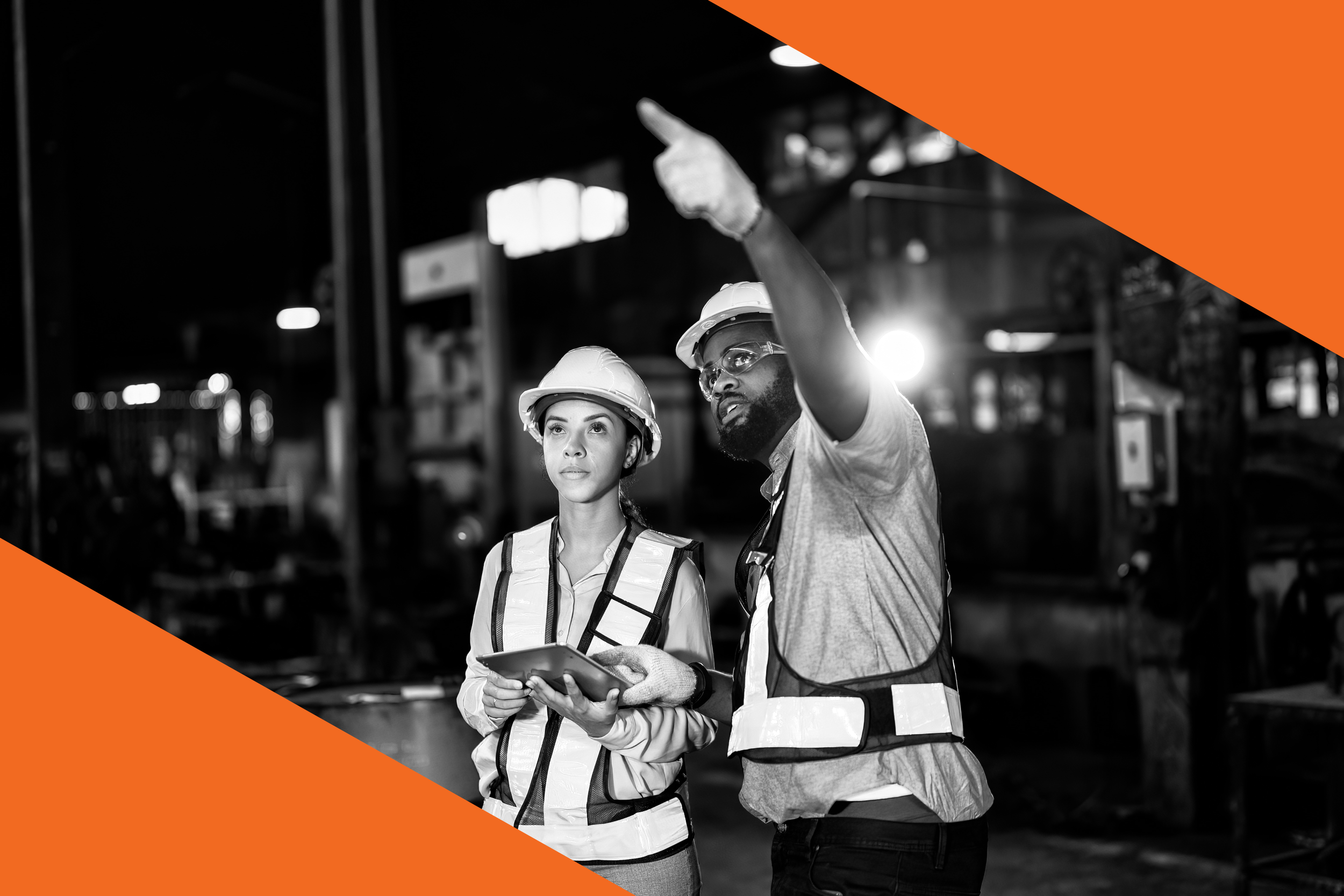 Zwei Bauarbeiter in Schutzhelmen und Sicherheitswesten unterhalten sich in einer Fabrikhalle, eine Person zeigt auf etwas und die andere hört aufmerksam zu.