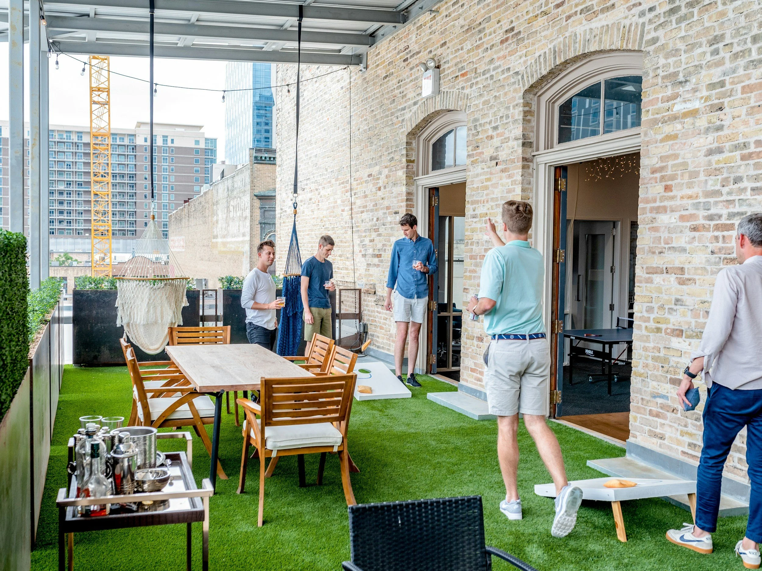 A group of men playing cornhole on a rooftop patio with artificial grass, a dining table, and city buildings in the background.